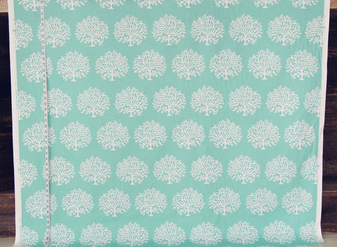 A roll of fabric with a repeating white tree or coral-like block print on a light aqua background, shown with a measuring tape along the left edge against a dark wood surface.