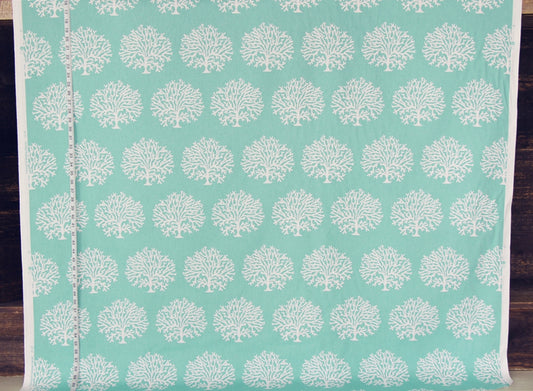 A roll of fabric with a repeating white tree or coral-like block print on a light aqua background, shown with a measuring tape along the left edge against a dark wood surface.