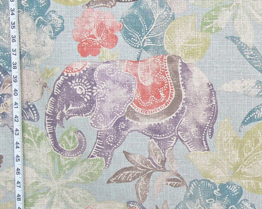 A close-up of a fabric pattern featuring a purple elephant with a red and brown saddle, surrounded by teal, green, and lavender leaves on a light blue background, with a white measuring tape visible on the left edge.