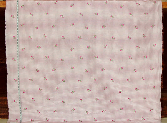 A square piece of light pink gingham fabric with a small repeating pattern of pink roses and green leaves, laid flat on a dark wooden surface.