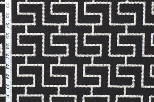 Black and white geometric patterned fabric with a repeating maze-like interlocking square design, shown close up with a white measurement ruler along the left edge.
