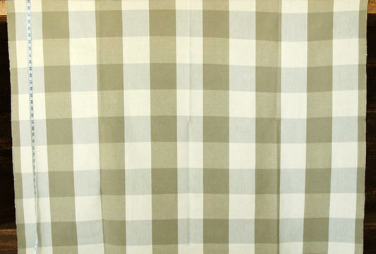 A close-up of a fabric with a gingham check pattern in sage green and off-white, laid flat on a dark wooden surface with a measuring tape visible on the left edge.