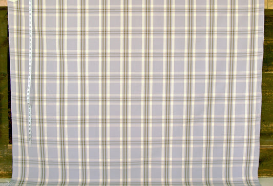 A close-up of a plaid fabric with a light gray base and intersecting cream, olive green, and tan stripes, shown flat with a measuring tape visible along the left edge.