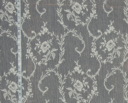White floral lace fabric with a repeating damask pattern, shown flat with a white measuring tape on the left side for scale.