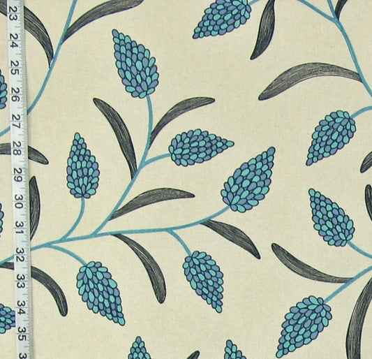 A close-up of a fabric or wallpaper pattern featuring blue-green floral buds and dark gray leaves on a warm cream background, with a white measuring tape visible along the left edge for scale.