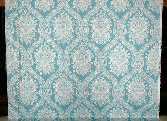 A close-up of a fabric or wallpaper sample with a repeating damask-style floral pattern in light aqua blue and white, shown against a dark background.