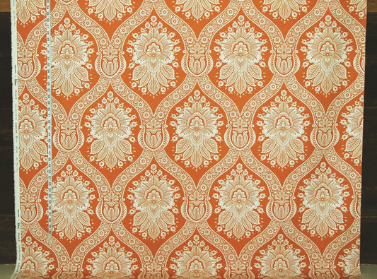 A close-up of patterned fabric with a repeating ornate damask-style floral motif in warm orange and off-white/cream.