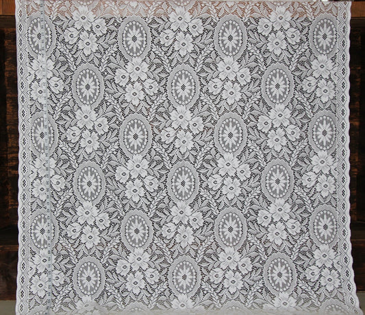 White floral lace textile with repeating oval medallion motifs and scalloped edges, laid flat against a dark wood background.