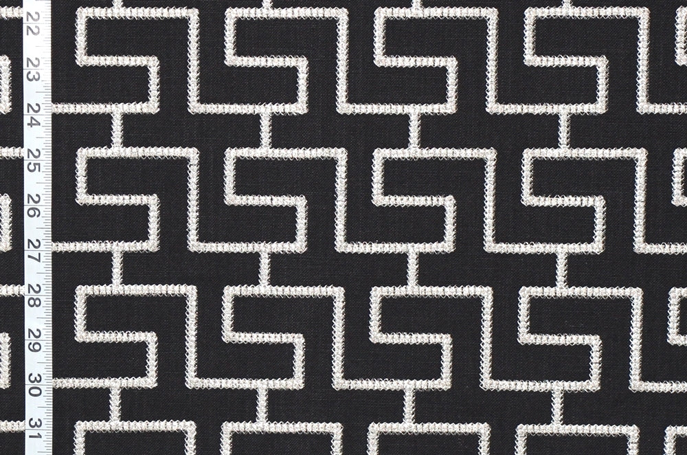 Black and white geometric patterned fabric with a repeating maze-like interlocking square design, shown close up with a white measurement ruler along the left edge.