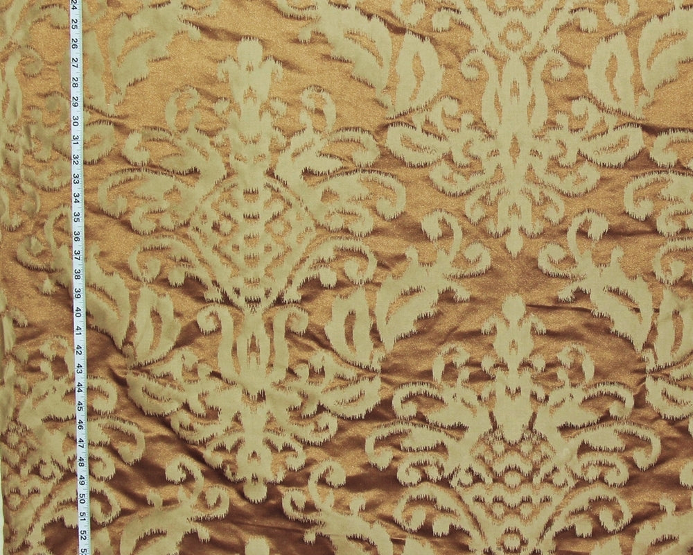 A close-up of damask patterned fabric in warm brown and beige, with a measuring tape visible along the left edge for scale.