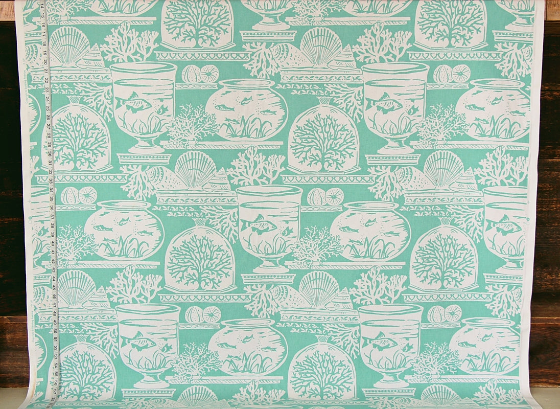 A roll of fabric or wallpaper featuring a repeating white toile-style pattern of fishbowl fish, shells, and coral motifs on a mint green background, shown against a dark wood surface.