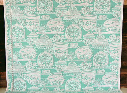 A roll of fabric or wallpaper featuring a repeating white toile-style pattern of fishbowl fish, shells, and coral motifs on a mint green background, shown against a dark wood surface.