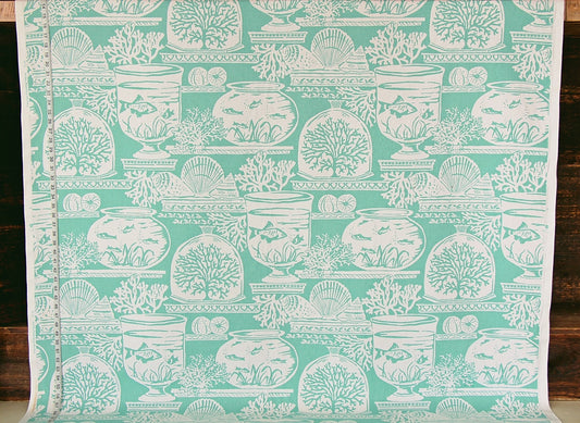 A roll of fabric or wallpaper featuring a repeating white toile-style pattern of fishbowl fish, shells, and coral motifs on a mint green background, shown against a dark wood surface.