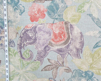 A close-up of a fabric pattern featuring a purple elephant with a red and brown saddle, surrounded by teal, green, and lavender leaves on a light blue background, with a white measuring tape visible on the left edge.