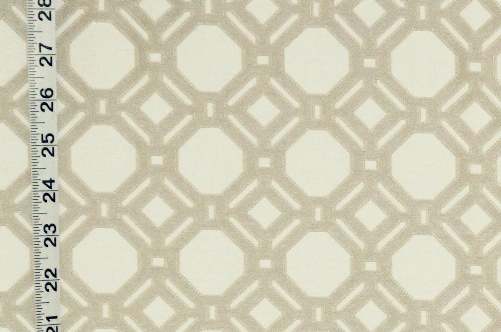 A close-up of a fabric or wallpaper pattern with a repeating geometric lattice design in warm beige over an off-white background, with a measuring ruler visible along the left edge for scale.