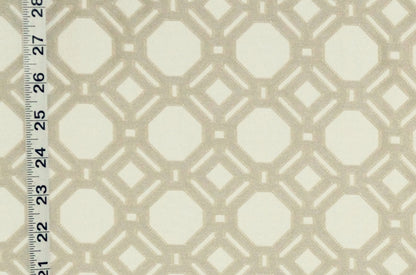 A close-up of a fabric or wallpaper pattern with a repeating geometric lattice design in warm beige over an off-white background, with a measuring ruler visible along the left edge for scale.