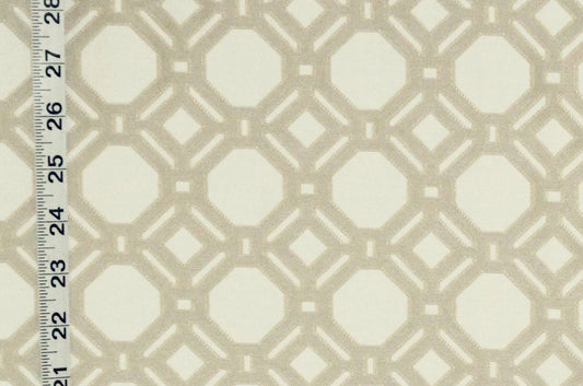 A close-up of a fabric or wallpaper pattern with a repeating geometric lattice design in warm beige over an off-white background, with a measuring ruler visible along the left edge for scale.