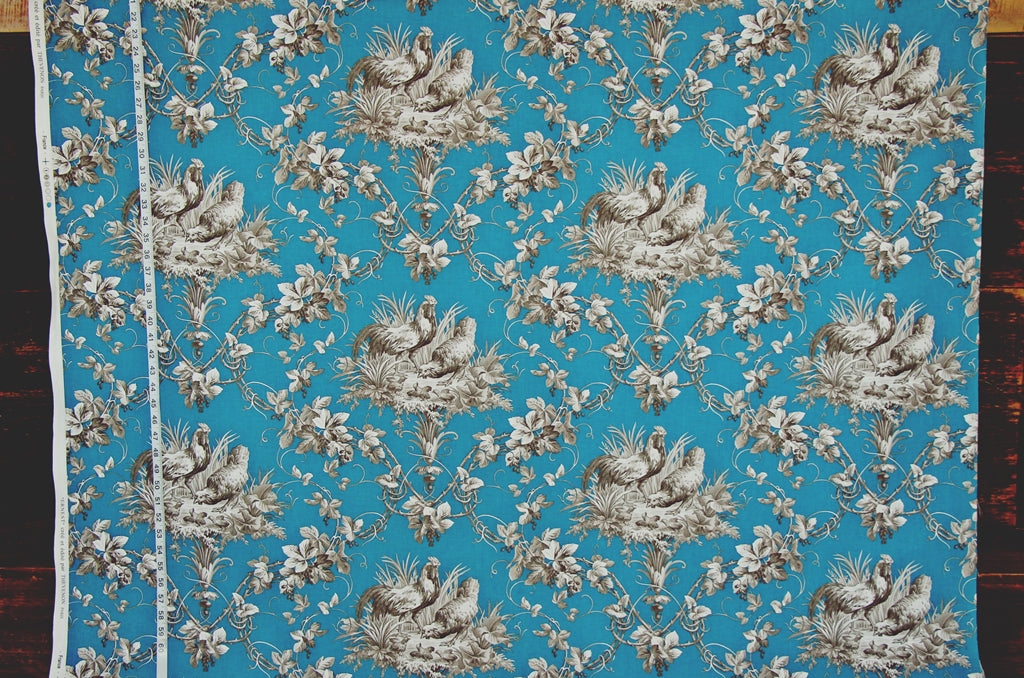 A square piece of fabric with a classic toile pattern featuring white birds and floral motifs on a bright turquoise blue background, laid flat with a white measuring tape visible along the left edge.