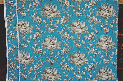 A square piece of fabric with a classic toile pattern featuring white birds and floral motifs on a bright turquoise blue background, laid flat with a white measuring tape visible along the left edge.
