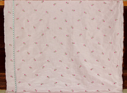 A square piece of light pink gingham fabric with a small repeating pattern of pink roses and green leaves, laid flat on a dark wooden surface.