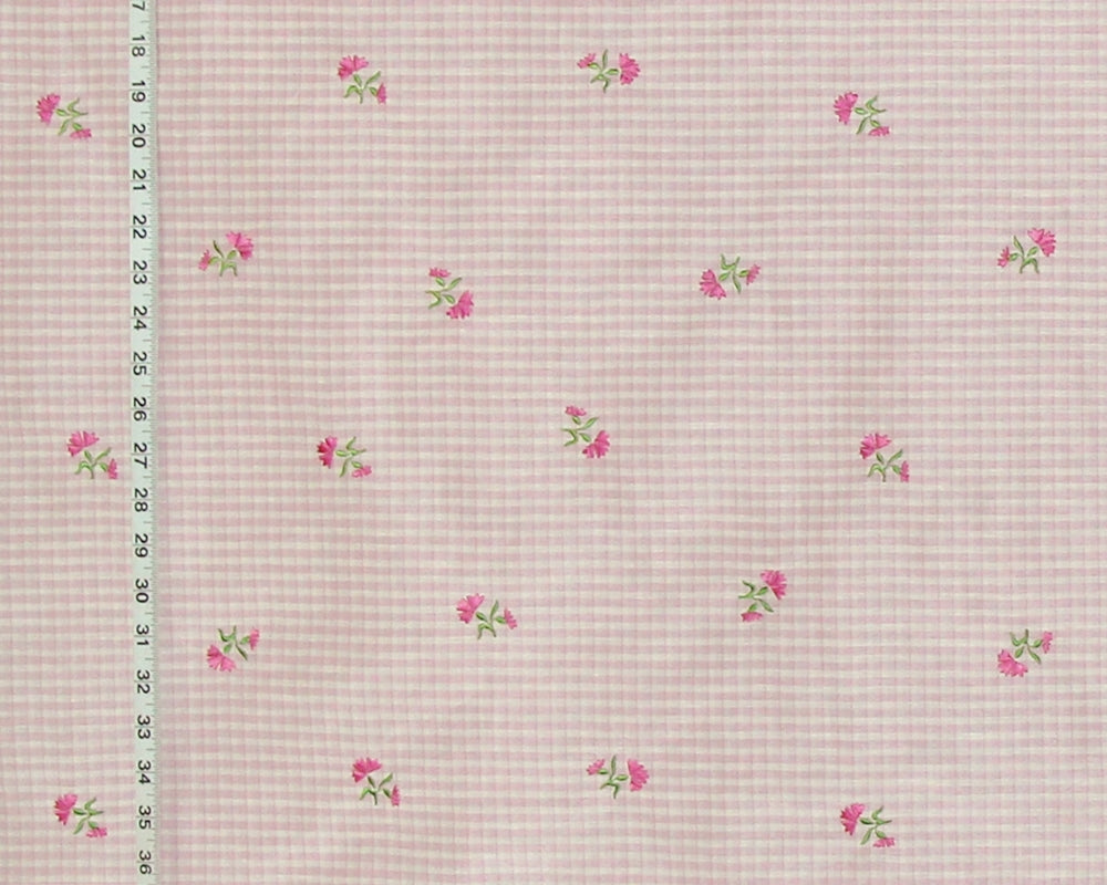 A close-up of a pink and white gingham fabric with a scattered pattern of small pink flowers and green leaves, shown with a measuring ruler on the left side.