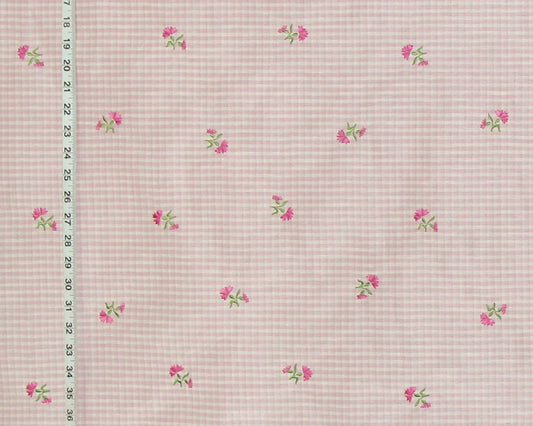A close-up of a pink and white gingham fabric with a scattered pattern of small pink flowers and green leaves, shown with a measuring ruler on the left side.