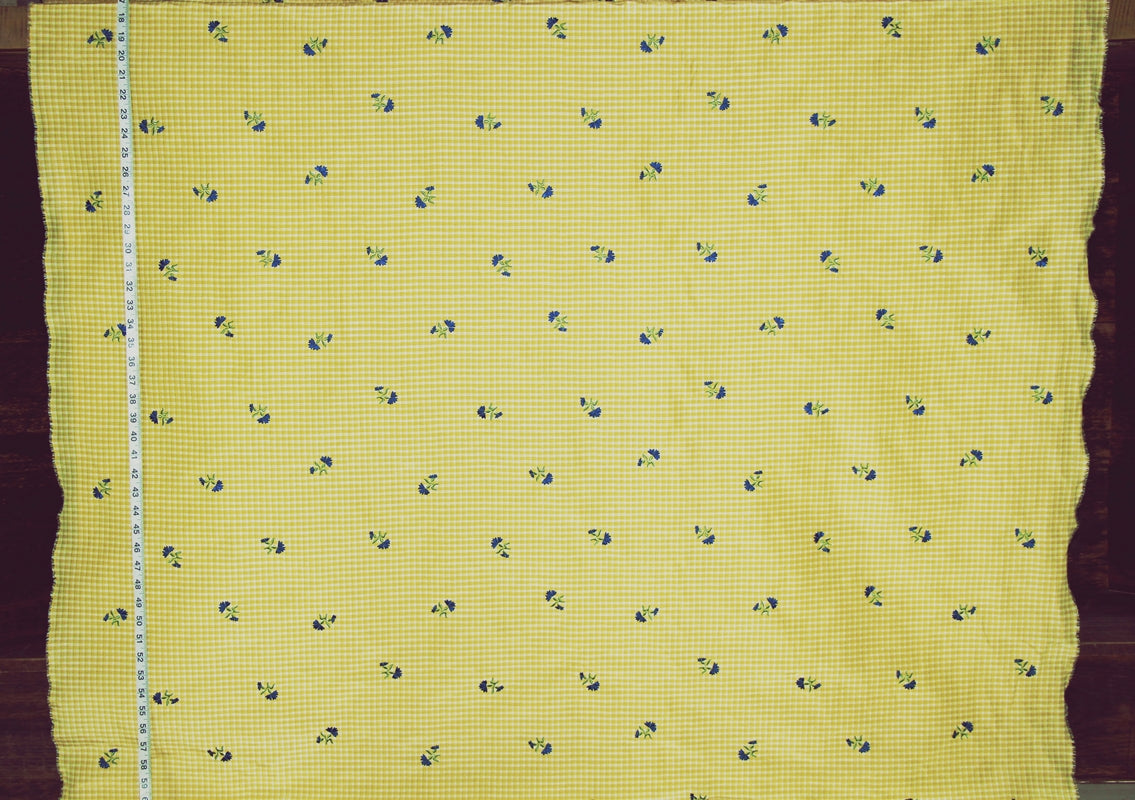 Yellow fabric with a small blue and white gingham check pattern and tiny blue floral motifs, shown flat with a measuring tape along one edge.