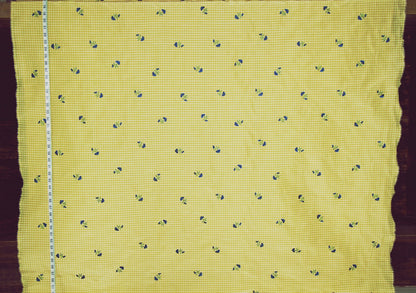 Yellow fabric with a small blue and white gingham check pattern and tiny blue floral motifs, shown flat with a measuring tape along one edge.