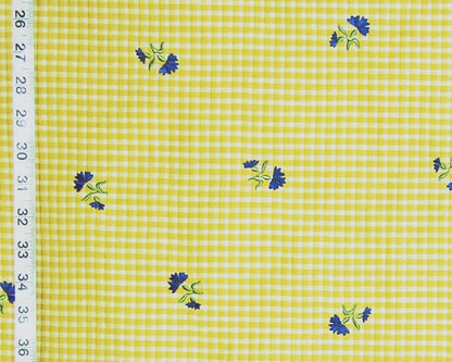Yellow and white gingham fabric with small blue and green floral motifs, shown with a measuring tape along the left edge.