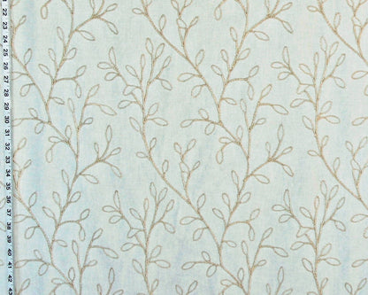 A close-up of a light blue fabric with a repeating pattern of delicate beige leafy vines, with a black measurement ruler visible along the left edge.
