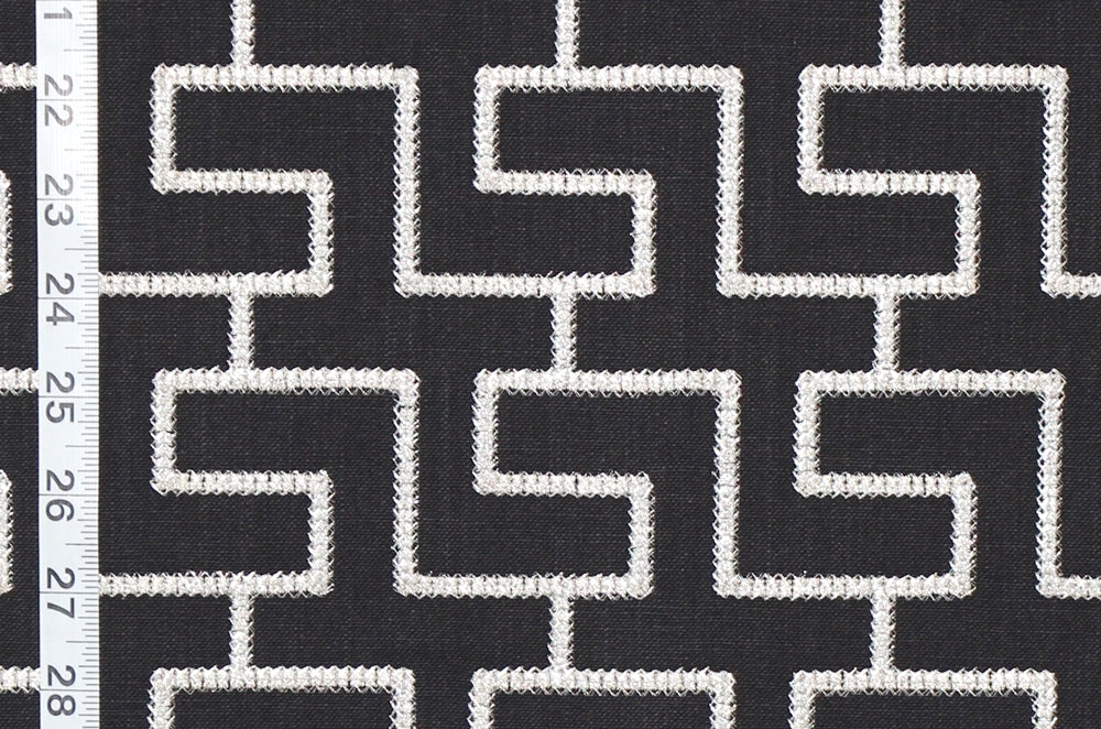 Black and white geometric patterned fabric with a repeating maze-like line design, shown close up with a white ruler on the left edge for scale.