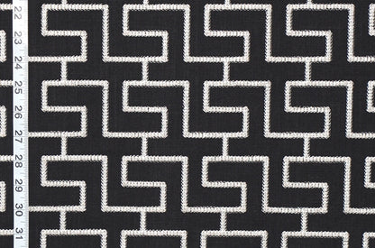 Black and white geometric patterned fabric with a repeating maze-like interlocking square design, shown close up with a white measurement ruler along the left edge.