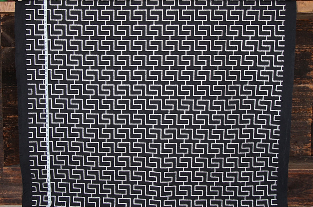 A black and white geometric patterned fabric, likely a tablecloth or runner, displayed flat against a rustic dark wood plank background.