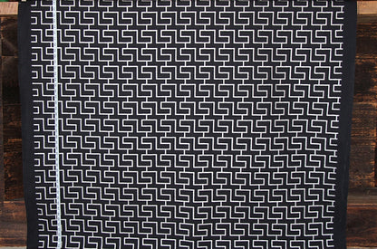 A black and white geometric patterned fabric, likely a tablecloth or runner, displayed flat against a rustic dark wood plank background.