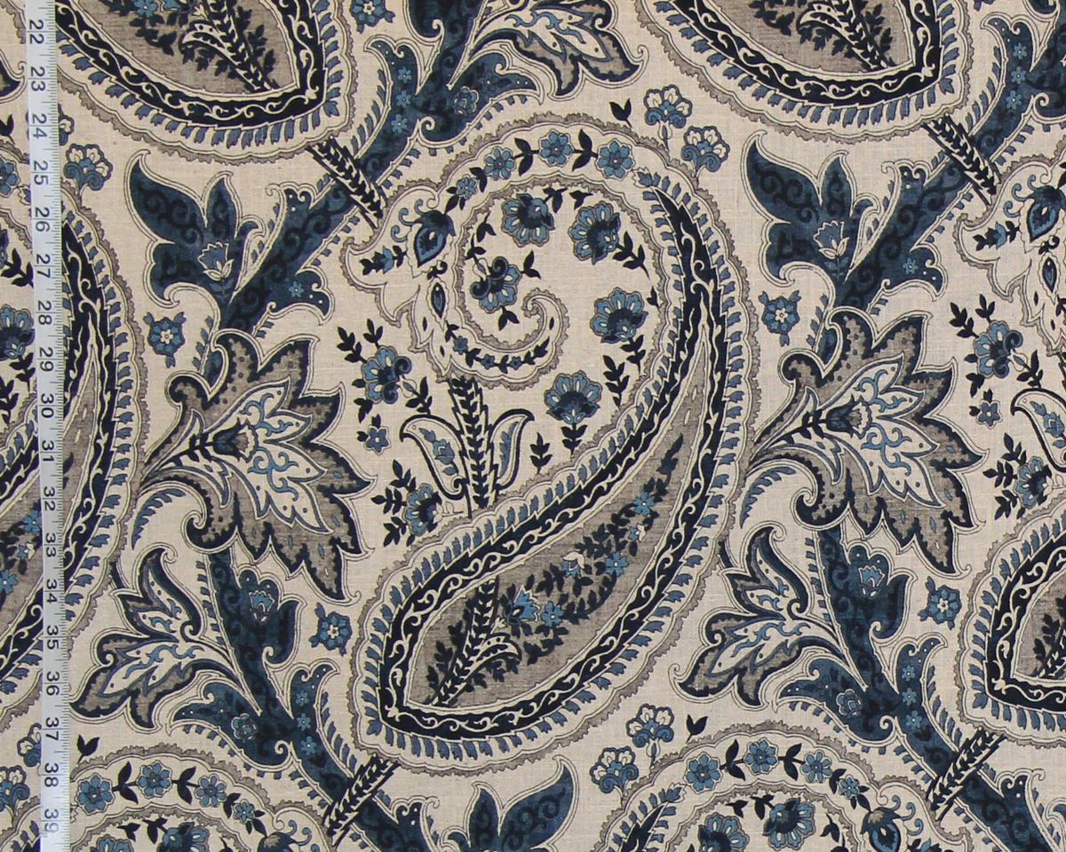 A close-up of a fabric pattern featuring a classic paisley design in navy blue, slate gray, and light blue on a warm beige background. A white fabric measuring tape is visible along the left edge to indicate scale.