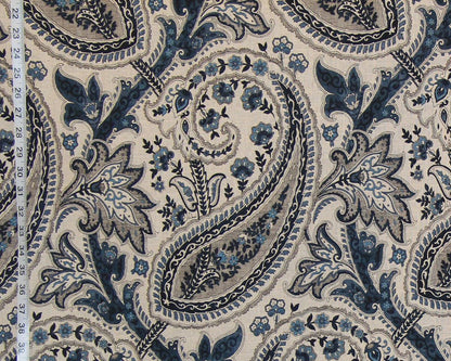 A close-up of a fabric pattern featuring a classic paisley design in navy blue, slate gray, and light blue on a warm beige background. A white fabric measuring tape is visible along the left edge to indicate scale.