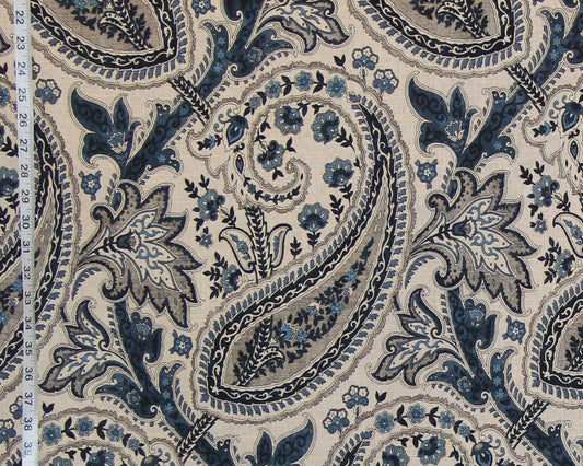 A close-up of a fabric pattern featuring a classic paisley design in navy blue, slate gray, and light blue on a warm beige background. A white fabric measuring tape is visible along the left edge to indicate scale.