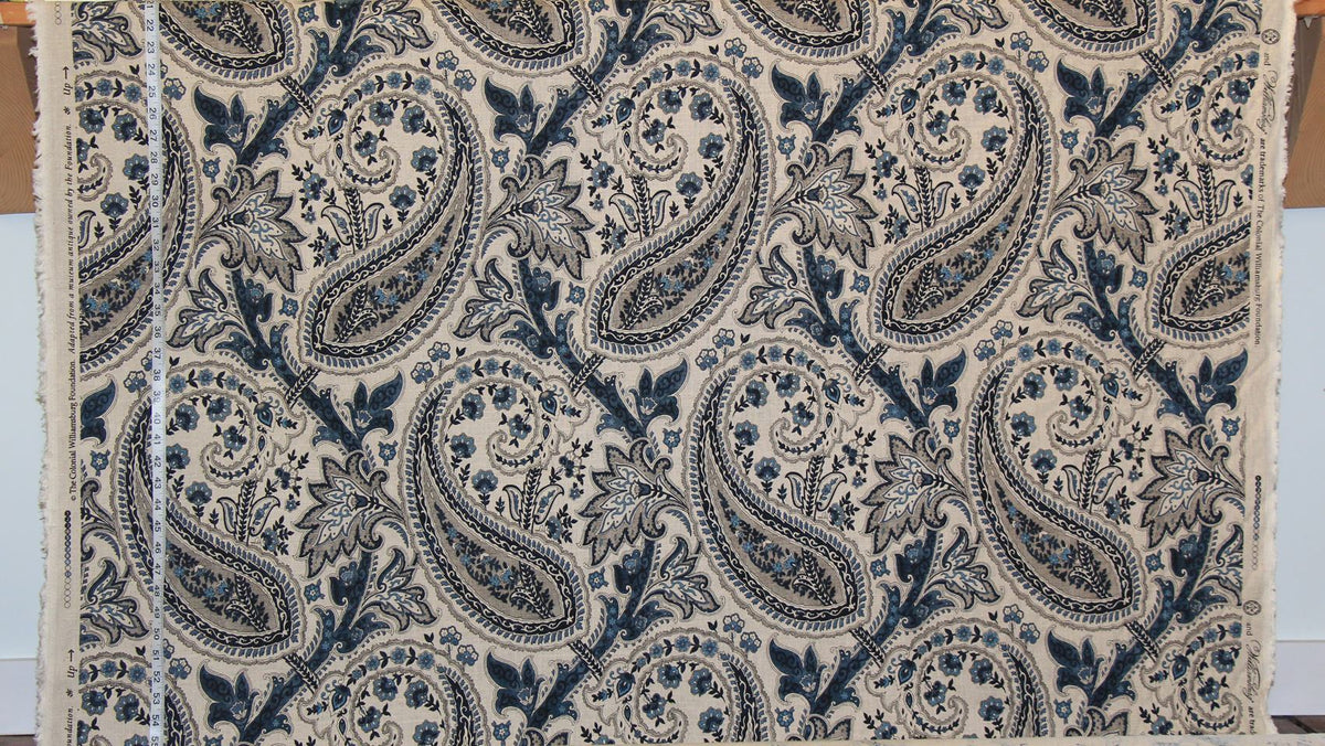 A close-up of a fabric or wallpaper sample featuring a classic navy blue and beige paisley pattern, with a white measuring tape visible along the left edge.