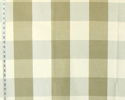 A close-up of a fabric surface with a large-scale gingham plaid pattern in muted olive green and off-white, with a measuring tape visible along the left edge.
