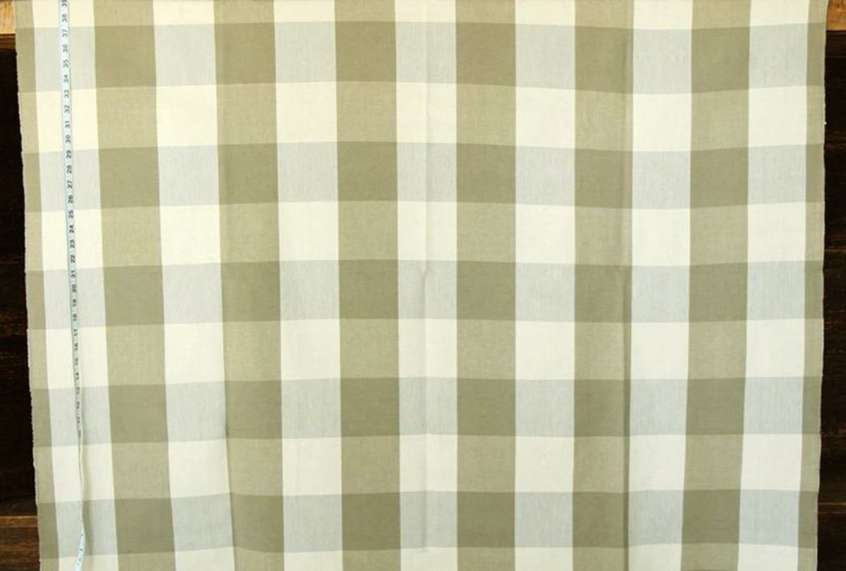 A close-up of a fabric with a gingham check pattern in sage green and off-white, laid flat on a dark wooden surface with a measuring tape visible on the left edge.