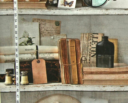 A styled vintage shelf scene with old books, a black bottle, small glass bottles, a wooden tag, and handwritten papers on a weathered gray shelf. A vertical measuring tape is visible along the left side, suggesting a scale or decor reference.