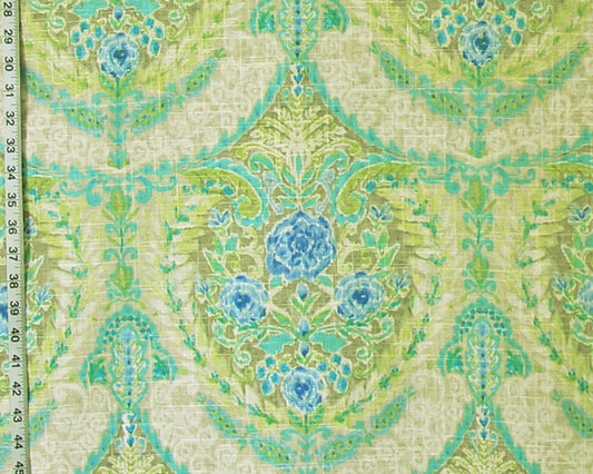 A close-up of a patterned fabric with a repeating damask-style floral design in light green, turquoise, and blue on a warm beige background. A measuring ruler is visible along the left edge to show scale.