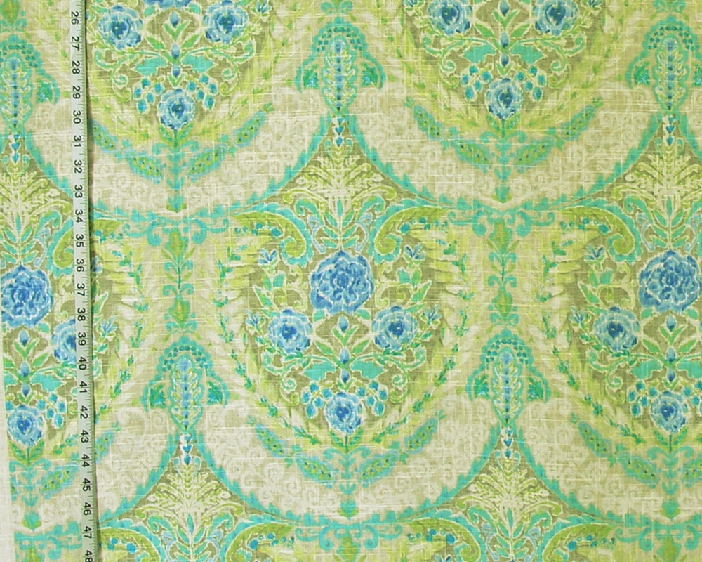 A close-up of patterned fabric with a repeating damask-style floral design in light green, aqua, turquoise, and blue on a warm cream background. A measuring tape is visible along the left edge for scale.