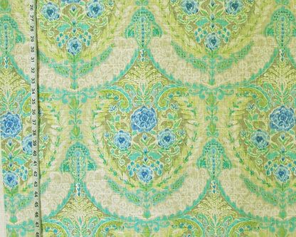 A close-up of patterned fabric with a repeating damask-style floral design in light green, aqua, turquoise, and blue on a warm cream background. A measuring tape is visible along the left edge for scale.