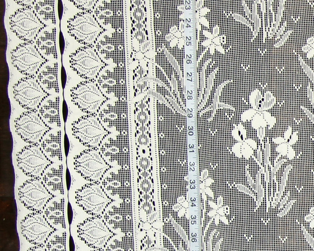 White floral lace fabric with multiple decorative borders and a measuring tape laid vertically to show scale.