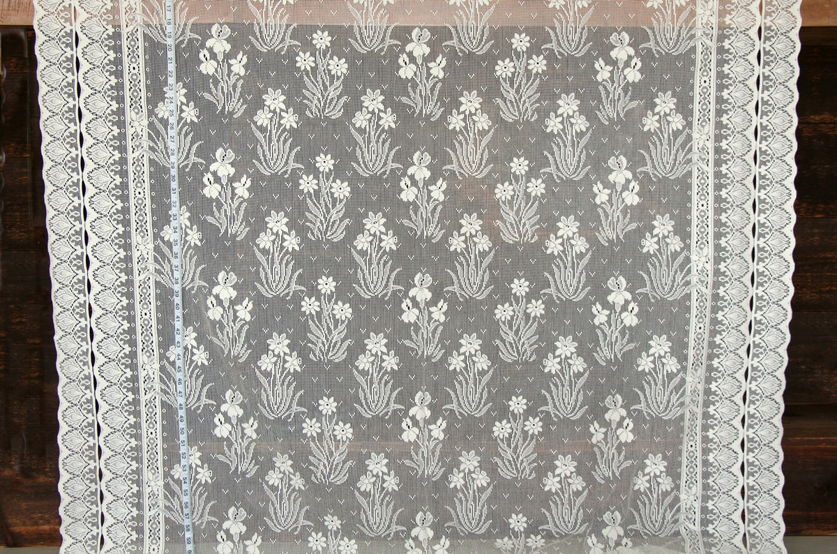 White floral lace tablecloth with a scalloped border, shown hanging against a dark wooden background.