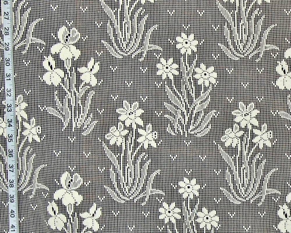 Gray and white floral lace fabric with a repeating pattern of iris-like flowers and leafy stems, shown close up with a measuring tape along the left edge.