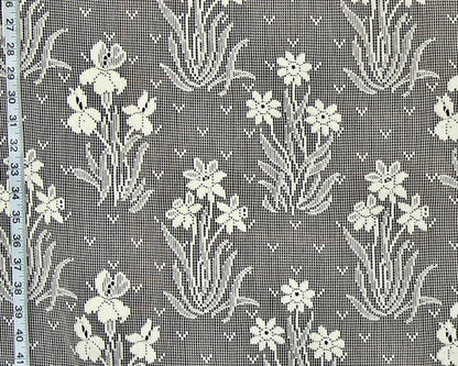 Gray and white floral lace fabric with a repeating pattern of iris-like flowers and leafy stems, shown close up with a measuring tape along the left edge.