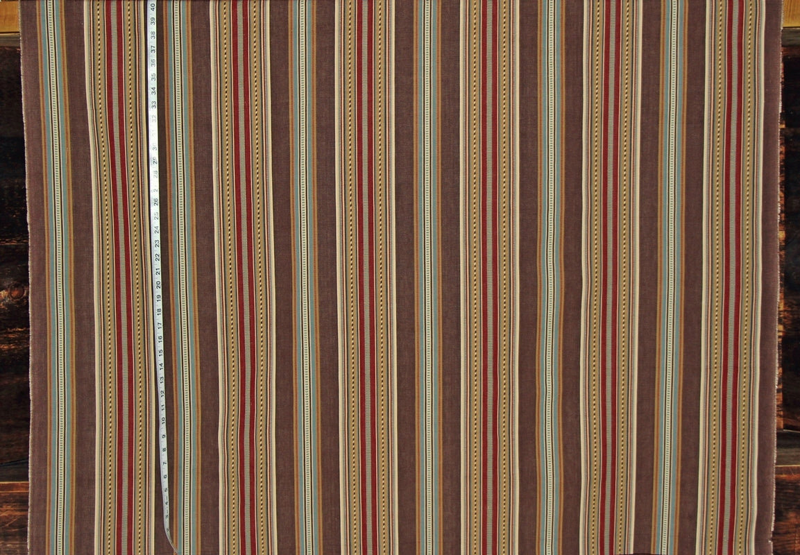 A close-up of a striped textile or fabric panel with vertical bands in brown, beige, red, and muted blue, with a measuring tape visible along the left side.