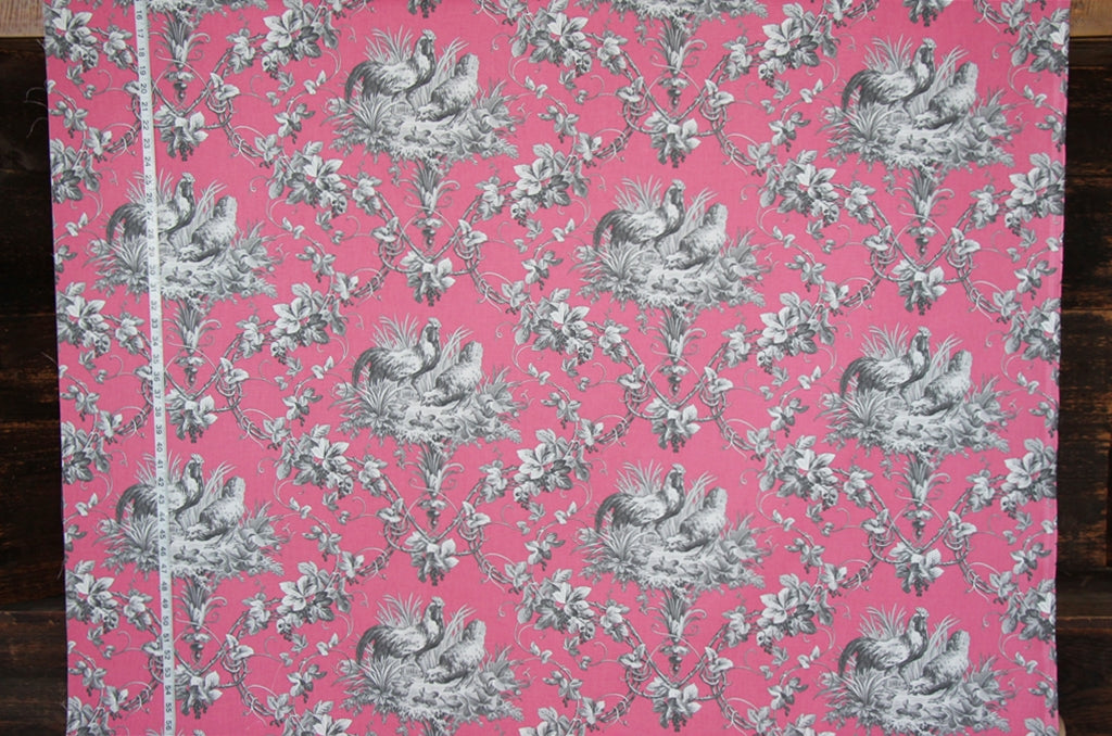 Pink fabric with a classic gray toile de Jouy pattern featuring peacocks, floral motifs, and a white measuring tape running vertically along the left edge, set against a dark wood background.